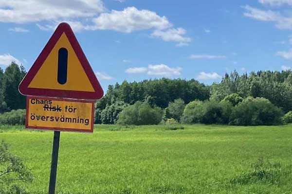 Bild på varningsskylt (trafikskylt) med texten Risk för översvämning där "Risk" är överstruket och ersatt med "Chans" - Chans till översvämning.