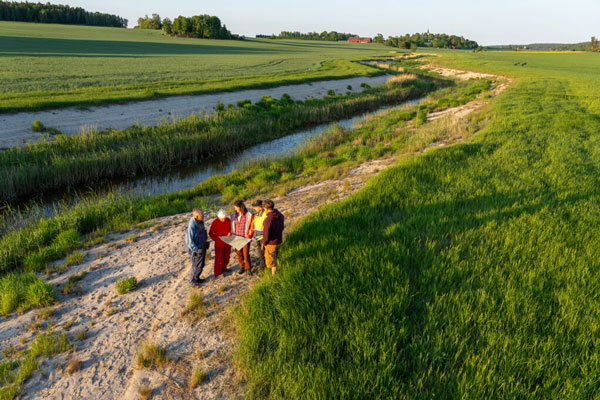 Fem personer står på stranden intill ett vattendrag som rinner genom ett jordbrukslandskap och tittar på en karta.